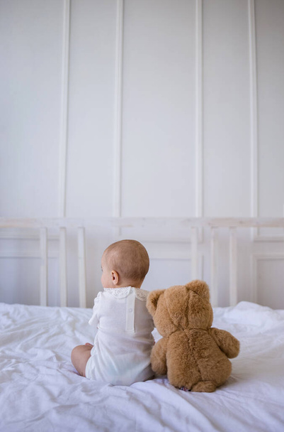 baby girl in a white bodysuit is sitting with her back with a teddy bear toy on a white cotton blanket on the bed in the room - Фото, зображення