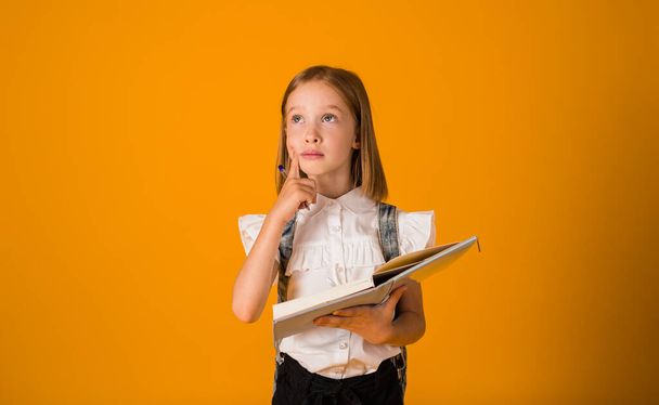 thoughtful schoolgirl in a uniform and with a backpack holds a notebook with a pen on a yellow background with a place for text - Valokuva, kuva