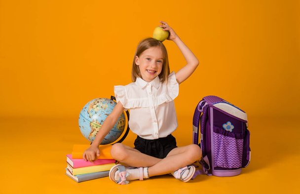 schoolgirl in uniform is sitting on the floor with school supplies and holding a green apple on a yellow background with a place for text on her head - Фото, изображение