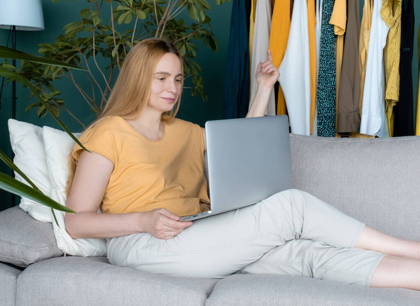 中年ブロンドの女性はソファに座って、ノートパソコンでオンラインで通信します。. - 写真・画像