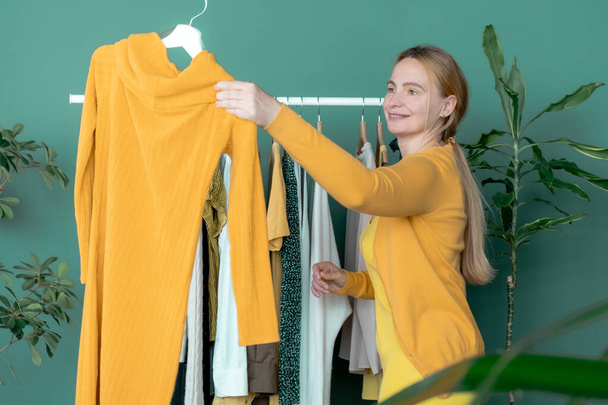 Średniowieczny Blondynka robi zakupy i patrzy na nową żółtą sukienkę w pobliżu półki z ubraniami - Zdjęcie, obraz