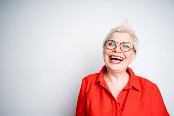 Cheerful elderly woman in glasses and in a red shirt laughs and smiles while looking at the camera - Foto, imagen