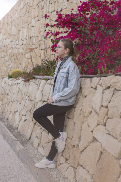 Full length portrait of a beautiful, young Caucasian sixteen year old pensive girl in a denim jacket posing outdoors near a park wall made of natural stone and bougainvillea flowers - Photo, Image