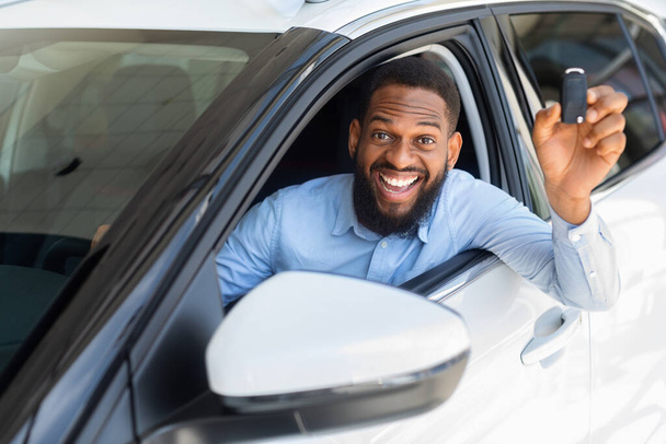 Szczęśliwy czarny człowiek siedzi w samochodzie, trzymając klucz i uśmiechając się do kamery - Zdjęcie, obraz