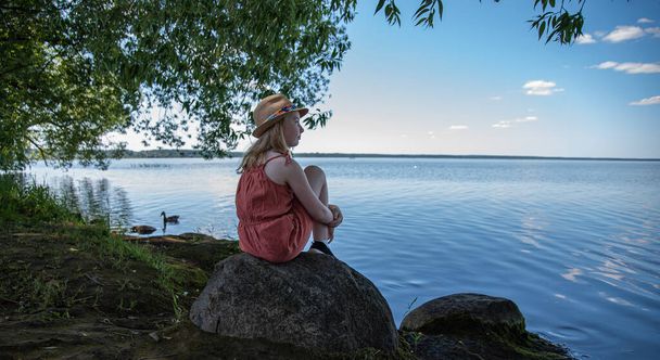 a little girl in an orange dress and hat is sitting on a stone on the shore of a lake  and looking dreamily into the distance on  a summer day - Photo, image