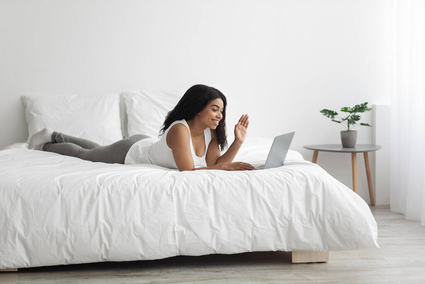 Jeune femme noire ayant appel vidéo sur ordinateur portable tout en étant couché sur le lit à la maison le week-end matin, copier l'espace - Photo, image