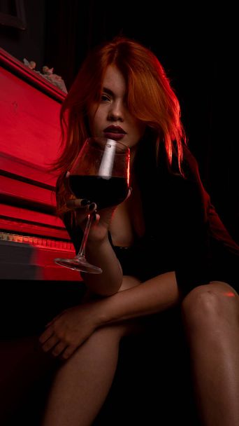 красивая молодая привлекательная женщина в вечернем платье позирует с винным бокалом и фортепиано  - Фото, изображение