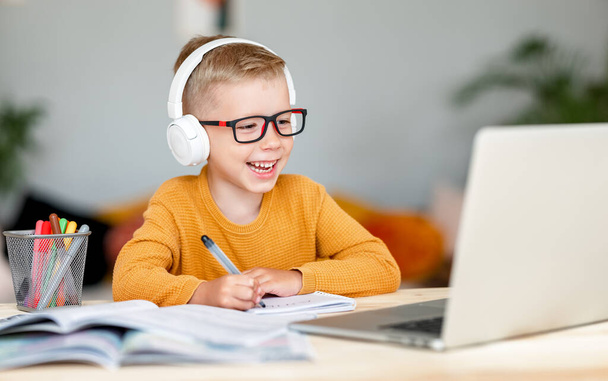 Ευτυχισμένο παιδί αγόρι με ακουστικά χαμογελώντας και κοιτάζοντας την οθόνη, ενώ κάνοντας βιντεοκλήση στο δάσκαλο κατά τη διάρκεια online μαθήματος στο σπίτι - Φωτογραφία, εικόνα