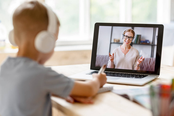 Kohdennettu lapsi poika kuulokkeissa katsoo huolellisesti opettajaa tehdessään videopuhelun online-oppitunnin aikana kotona - Valokuva, kuva