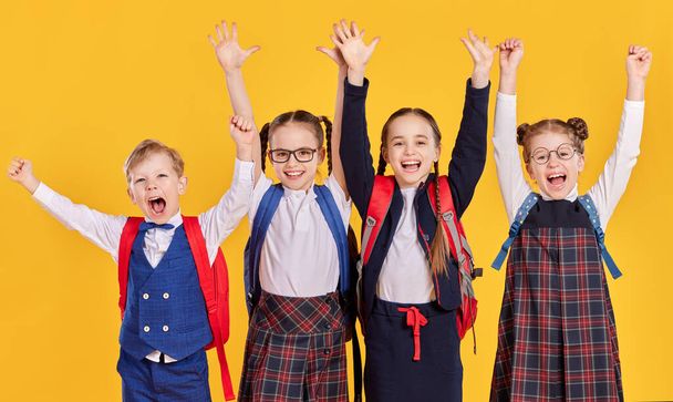Skupina nadšených dětí základních spolužáků ve školní uniformě s batohem zvedat ruce a dívat se na kameru s vzrušením proti žlutému pozadí - Fotografie, Obrázek