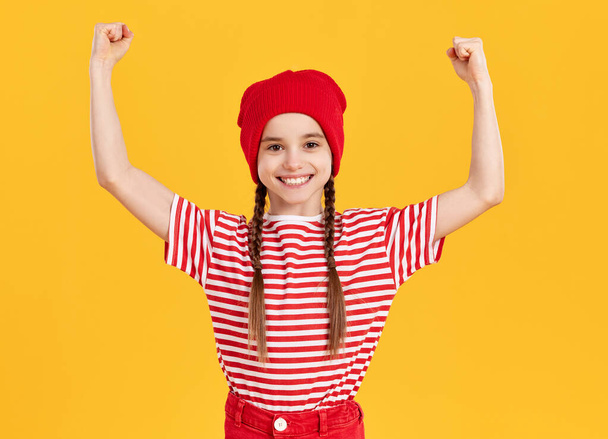 Joyeux hipster preteen fille portant chapeau rouge et chemise rayée levant les bras tout en célébrant la réalisation sur fond jaune vif - Photo, image