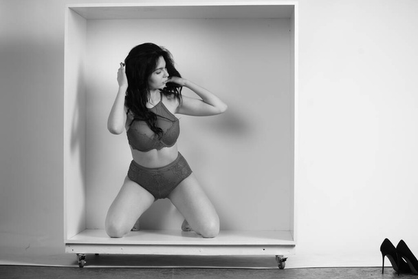 bella donna bruna che indossa lingerie posa in cubo bianco in studio  - Foto, immagini