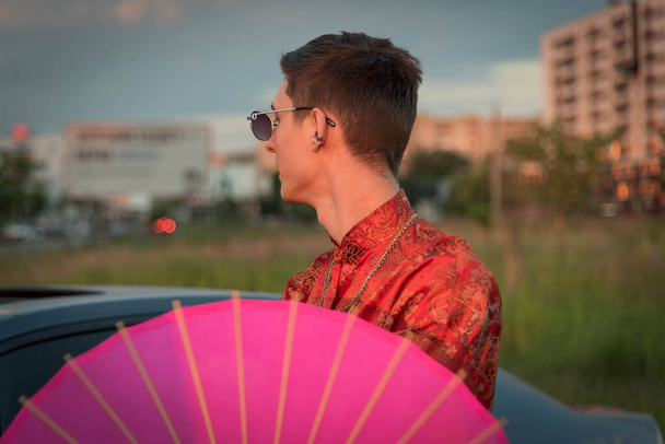 Positiivinen mies teini nokkela pukeutunut katu tyyli vaatteita punainen kimono ketjuilla kaulassa vaaleanpunainen sateenvarjo. Nuoruuden ja elämäntavan käsite. Ylellinen rap-artisti. - Valokuva, kuva
