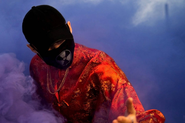 Positive male teenager wit dressed in street style clothes red kimono with chains around neck, black cap and black medical mask . Youth and lifestyle concept. Luxury rap artist in violet smoke. - Photo, image