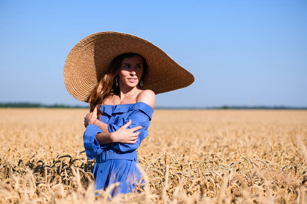 Menina sorridente em vestido vintage romântico azul e chapéu de aba larga posando no campo de trigo de verão, aparência de moda rural. Mulher abraça-se. - Foto, Imagem