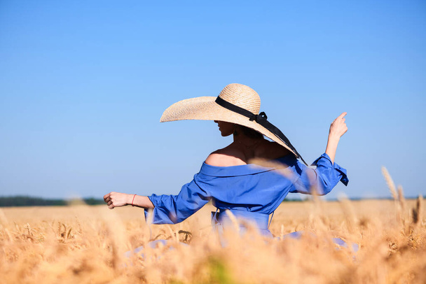Дівчинка залишається у широкому крихітному капелюсі та синій сукні всередині літнього пшеничного поля, витонченої моди. Жінка танцює серед вух
. - Фото, зображення