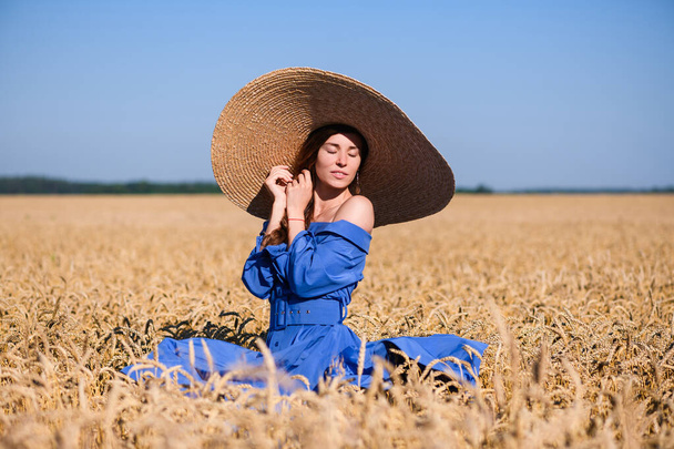 Romantic vintage lady in blue midi dress with belt and wide brimmed hat stand inside countryside golden wheat field - Foto, afbeelding