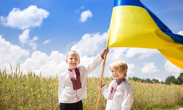 Zwei fröhlich lächelnde Jungen in ukrainischen Nationaltrikots halten vor dem Hintergrund eines Weizenfeldes eine große gelb-blaue Fahne hoch. Ukrainische Staatsbürgerschaft. Patriotische Erziehung. Unabhängigkeitstag - Foto, Bild