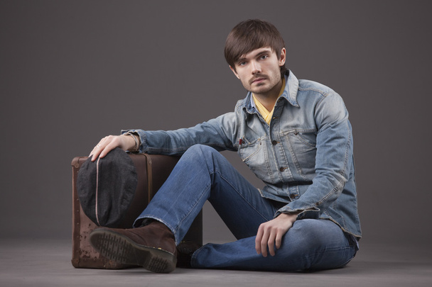 Мужчина модели в джинсовой одежде сидит на земле со старым чемоданом
 - Фото, изображение