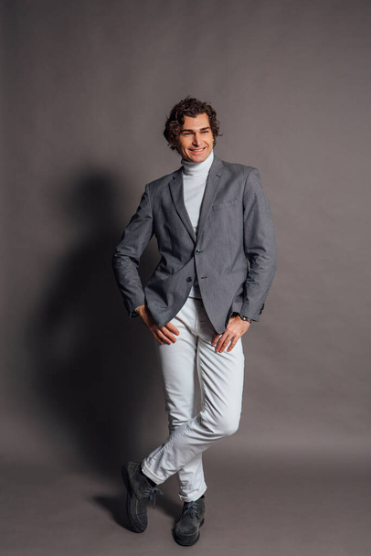 Fashion portrait of a tall handsome man dressed in white turtleneck, white jeans and grey jacket posing on the grey background - Φωτογραφία, εικόνα