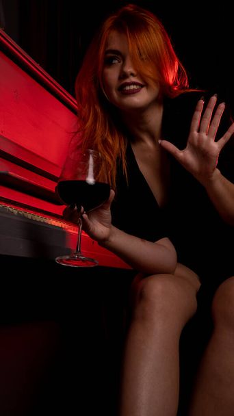Akşam elbisesi içinde şarap kadehi ve piyanoyla poz veren güzel ve çekici bir kadın.  - Fotoğraf, Görsel