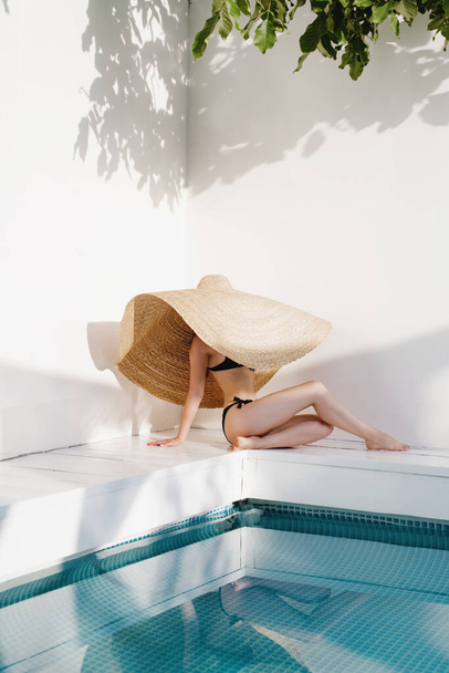 Unrecognizable woman in black bikini and big hat relaxing near swimming pool. - Foto, Imagen
