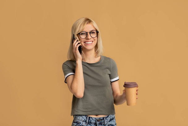 Красивая молодая леди пьет кофе на вынос и разговаривает по мобильному телефону на фоне красочной студии - Фото, изображение