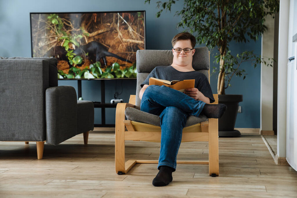 Mladý muž s Downovým syndromem čtení knihy, zatímco sedí v křesle doma - Fotografie, Obrázek