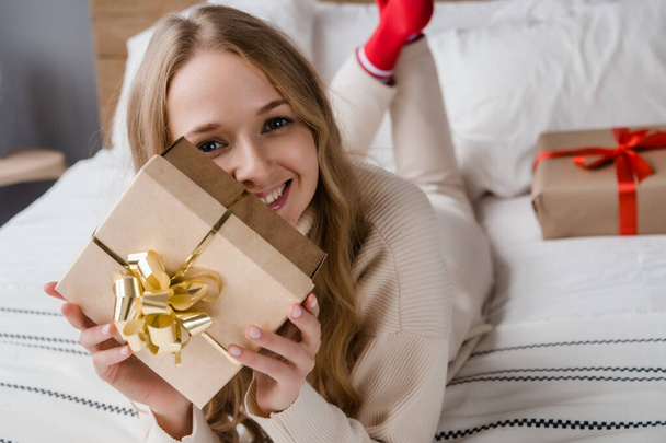 Śliczna uśmiechnięta kobieta z pudełkiem prezentów leżąca w łóżku i patrząca w kamerę. Koncepcja święta Bożego Narodzenia lub urodziny - Zdjęcie, obraz