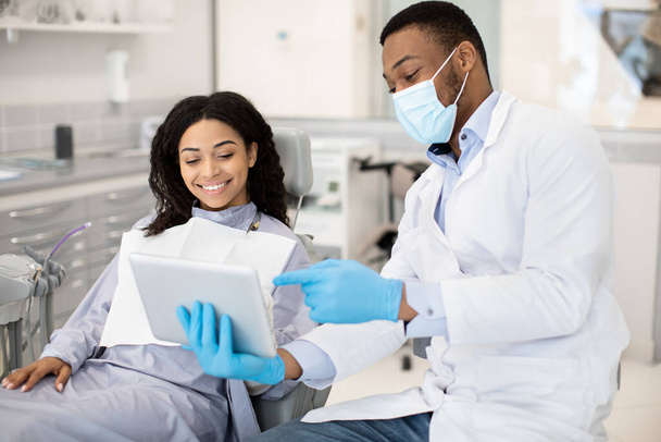 Zwarte tandarts met digitale tablet consulting vrouwelijke patiënt in de moderne kliniek - Foto, afbeelding