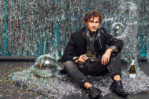 Tall attractive man sitting on the floor over the sparkle background and mirror disco ball holding glass of champagne. Party concept. - Foto, Bild