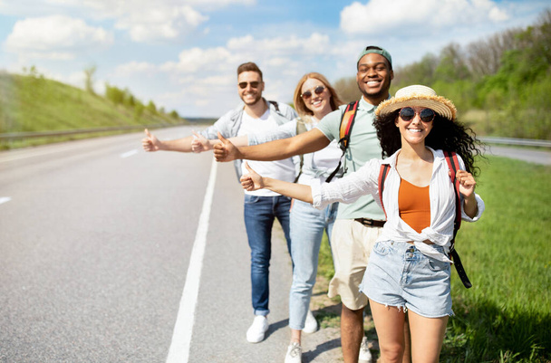Молодые многорасовые друзья стоят на шоссе, показывая автостопом жест, останавливая машину, отправляясь в путешествие вместе - Фото, изображение