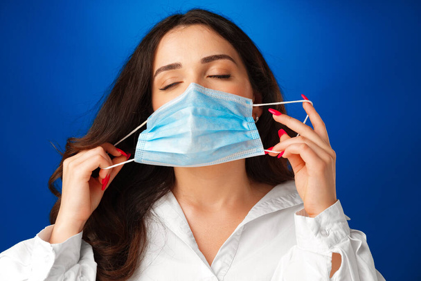 Young woman takes medical mask off her face against blue background - Valokuva, kuva