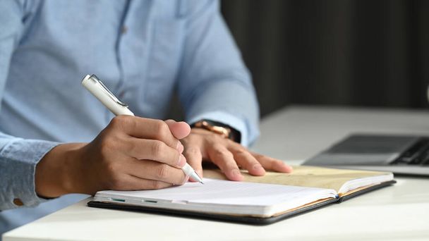 Zamknij widok biznesmena trzymającego długopis podczas pisania w notatniku i siedząc w miejscu pracy. - Zdjęcie, obraz