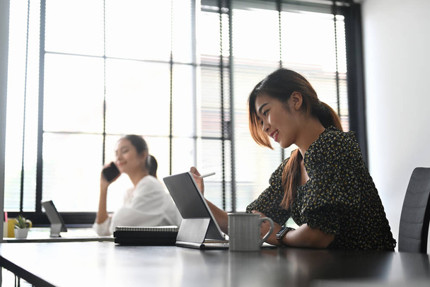 Due donne d'affari felici sono colleghi seduti insieme a un tavolo in un ufficio moderno e lavorano su tablet per computer. - Foto, immagini