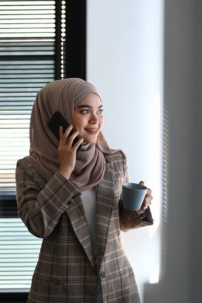Portret van een jonge vrolijke moslim vrouw in hijab praat op mobiele telefoon en houdt koffiebeker op kantoor. - Foto, afbeelding