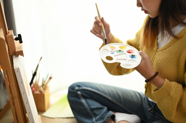 Cropped shot of young female artist with palette in her hand painting on canvas while sitting on floor. - Photo, Image