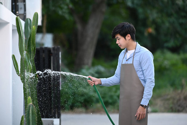 A young man is holding green rubber hose and watering plants at home. Hobby lifestyle concept. - Photo, Image