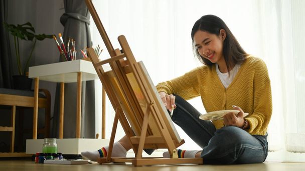 A young female artist in yellow sweater with palette in her hand is painting on canvas on floor. - Photo, Image