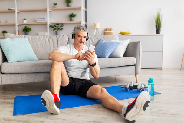 Thuistraining. Happy atletische volwassen man luisteren muziek in hoofdtelefoon tijdens het zitten op de mat en met behulp van smartphone - Foto, afbeelding