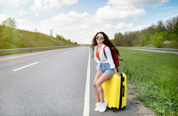 Красивая молодая женщина сидит на чемодане вдоль дороги, машет вниз автомобиль, автостопом на обочине дороги, имея летнее путешествие - Фото, изображение