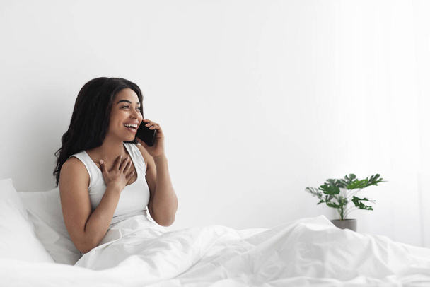Happy african american lady sitting on bed and talking on smartphone, looking at copy space, resting in morning at home - Photo, image