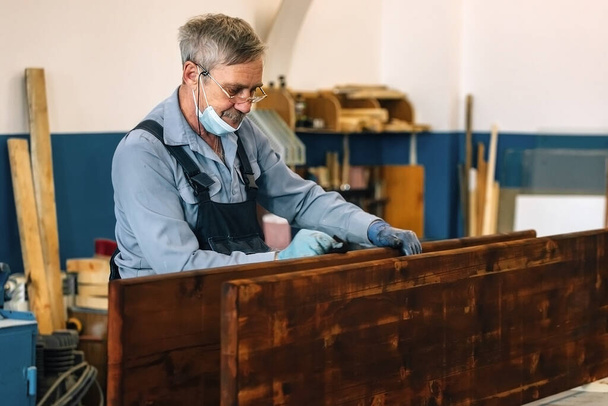 Ένας ηλικιωμένος ζωγραφίζει ξύλινες σανίδες με σκούρο βερνίκι σε εργαστήριο ξυλουργικής. Συνταξιούχος με ιατρική μάσκα  - Φωτογραφία, εικόνα