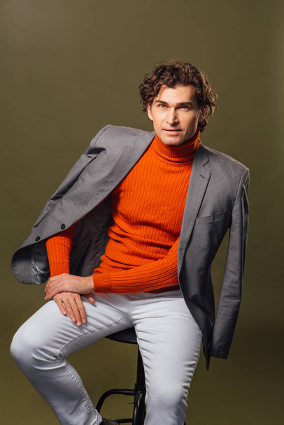 Portrait of a tall handsome man dressed in orange turtleneck, grey jacket and white jeans posing on the green background - Фото, зображення