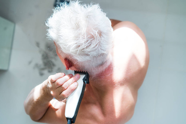 Top view white hair man doing self haircut with a clipper and looks in the mirror. Self-care at home in the conditions of global quarantine and closed hairdressers and beauty salons. electric razor - Photo, Image