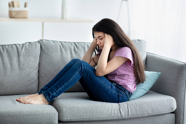 Unhappy teen girl holding her head in despair, feeling depressed, sitting on sofa at home, copy space - Photo, Image