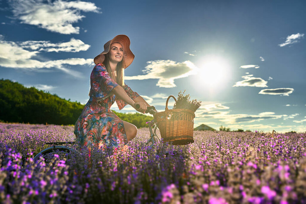 Beautiful young woman riding her bicycle in a lavender field at sunset - Фото, изображение