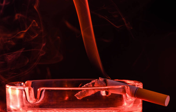 Red smoke rising from burning cigarette in glass ashtray maroon, cigarette butt - Foto, Bild