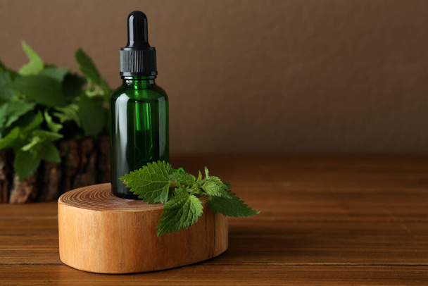 Glass bottle of nettle oil with dropper and leaves on wooden table against brown background, space for text - Foto, imagen