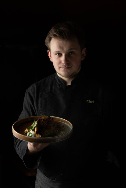 Un cocinero con chaqueta de chef negra sirve un plato cocinado en un plato - Foto, Imagen
